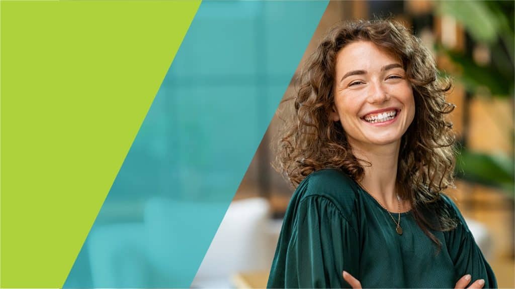 Une femme souriante, portant une blouse verte, se tient debout dans un bureau moderne, les bras croisés.