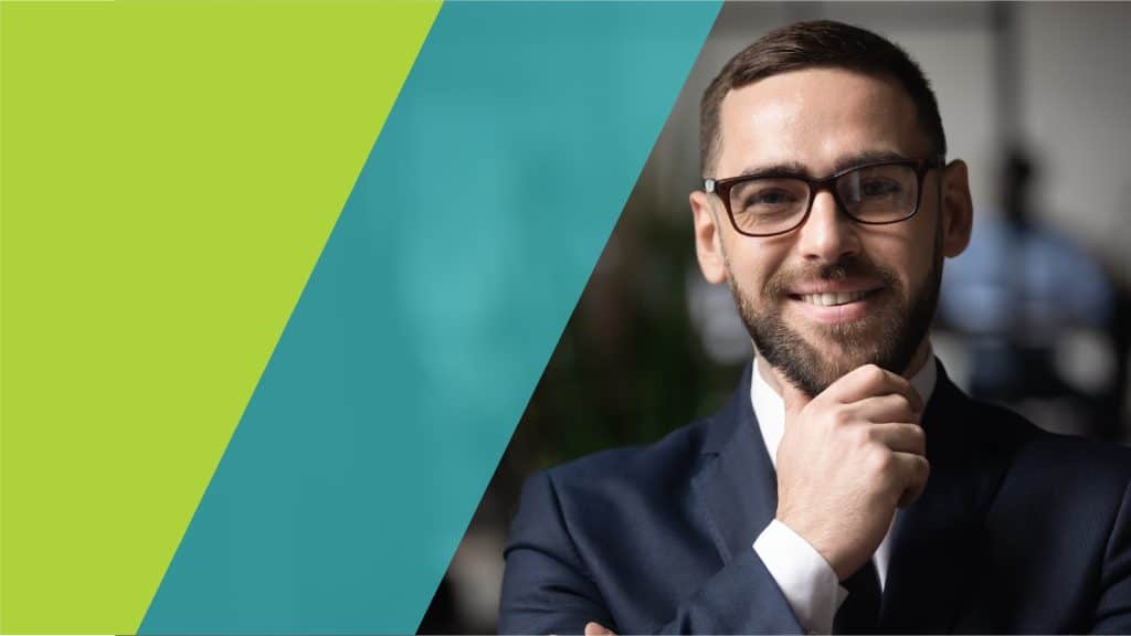 Un homme en costume souriant, avec des lunettes, posant avec assurance dans un bureau moderne.