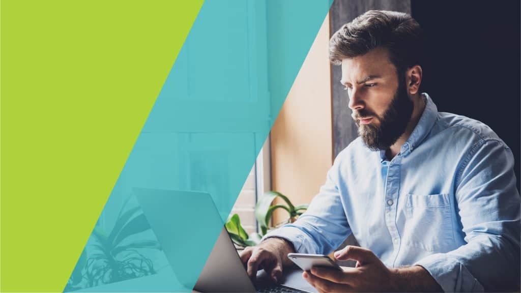 Un homme concentré travaille sur un ordinateur portable tout en tenant un smartphone, dans un environnement lumineux et calme.