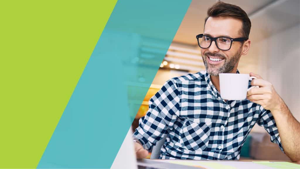 Un homme souriant portant des lunettes, assis devant un ordinateur portable, tenant une tasse de café.