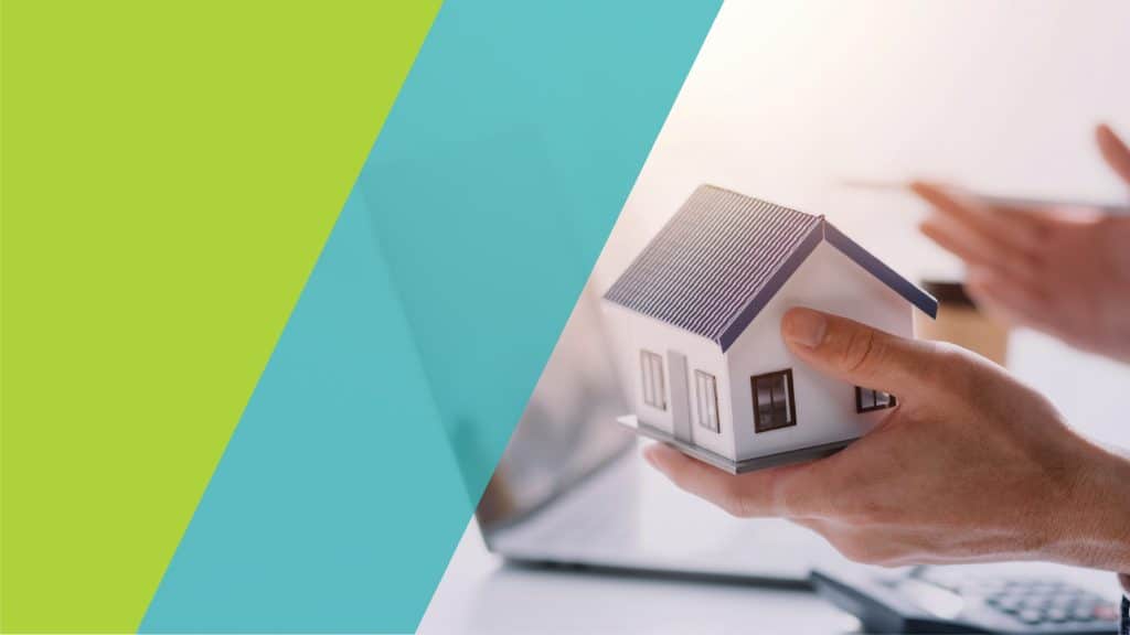 Un modèle réduit de maison dans une main, avec un ordinateur portable et des documents en arrière-plan.