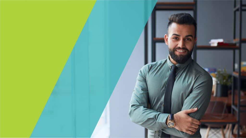 Un homme souriant, habillé de manière professionnelle, croise les bras dans un bureau moderne.
